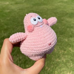 May include: A handmade pink crochet Patrick Star keychain. The plush figure has large white eyes with black pupils, rosy cheeks, and a small smile. The keychain is held in a hand against a blurred green background.