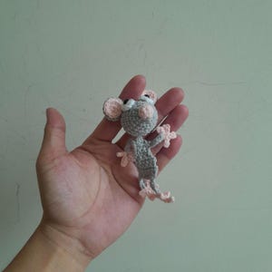 Könnte beinhalten: Eine kleine, gehäkelte Mausfigur in Grau mit rosa Akzenten an Ohren, Pfoten und Füßen. Die Maus hat schwarze Augen. Die Figur wird in einer Hand gehalten, um die handgefertigten Details und die Größe zu zeigen.