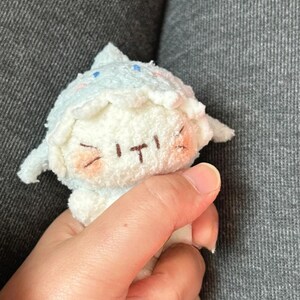 Peut inclure: Une petite poup&eacute;e en peluche aux couleurs bleu clair et blanc. La poup&eacute;e a un chapeau pointu, des joues roses et un visage cousu avec une expression souriante. La poup&eacute;e est tenue dans une main.