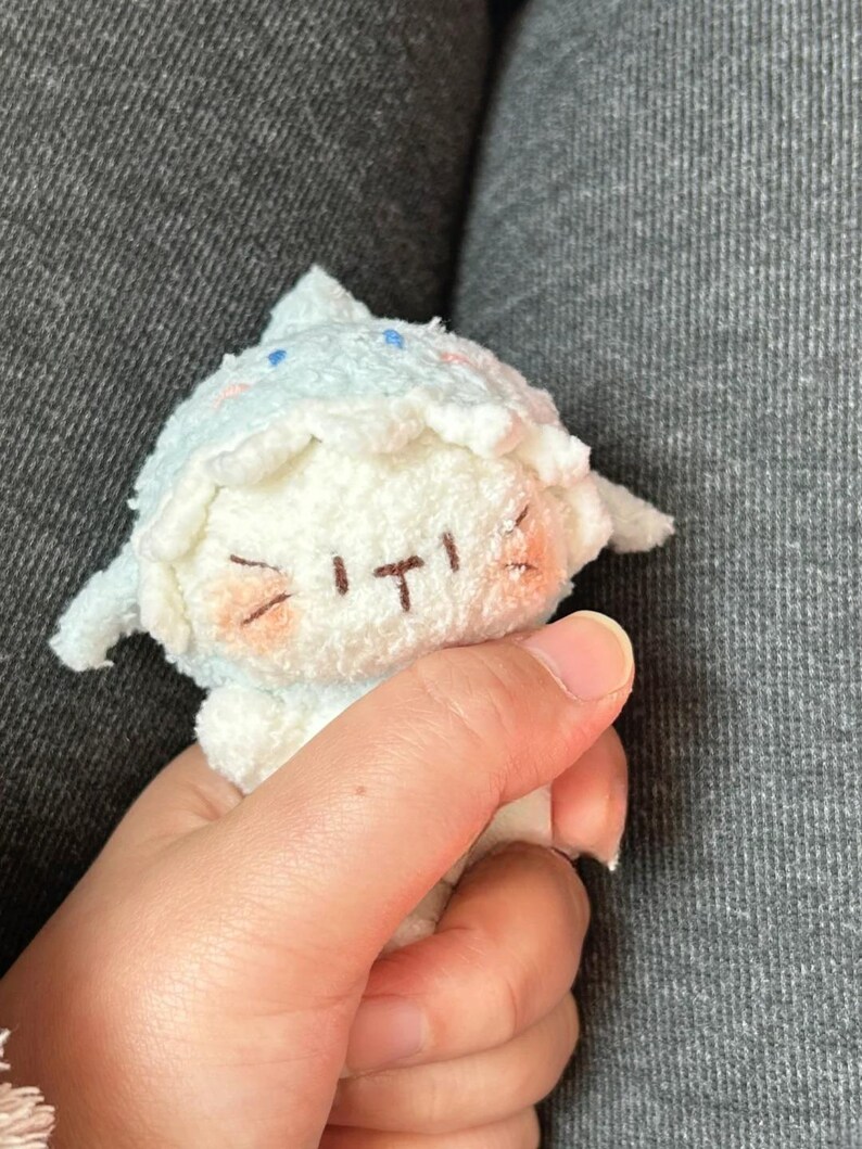 Peut inclure: Une petite poup&eacute;e en peluche aux couleurs bleu clair et blanc. La poup&eacute;e a un chapeau pointu, des joues roses et un visage cousu avec une expression souriante. La poup&eacute;e est tenue dans une main.