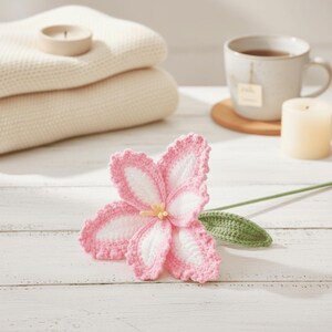 Op de afbeelding: Een handgemaakte gehaakte bloem met roze en witte bloemblaadjes, een gele kern en een groene stengel en blad. De bloem ligt op een wit houten oppervlak, met crèmekleurige dekens, een theekopje en een kaars op de achtergrond.