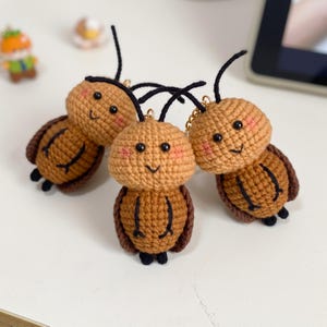 May include: Three handmade crochet keychains shaped like small brown insects. Each figure features a round head with black bead eyes, a stitched smile, pink blush, and two black antennae. The bodies are textured with brown yarn and dark brown wings.