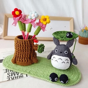 Könnte beinhalten: Ein handgefertigter Häkel-Stifthalter in Form eines Baumstumpfes, der Stifte mit bunten gehäkelten Blumen hält. Eine Häkel-Totoro-Figur und zwei schwarze Häkeltiere sitzen auf einer grünen, blattförmigen Häkelmatte. Ein skurriles Schreibtischzubehör.