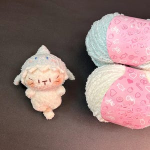 Peut inclure: Une peluche blanche avec un chapeau et des d&eacute;tails bleu clair, &agrave; c&ocirc;t&eacute; de deux pelotes de laine blanche et bleu clair. La laine a des &eacute;tiquettes roses avec de petites illustrations.