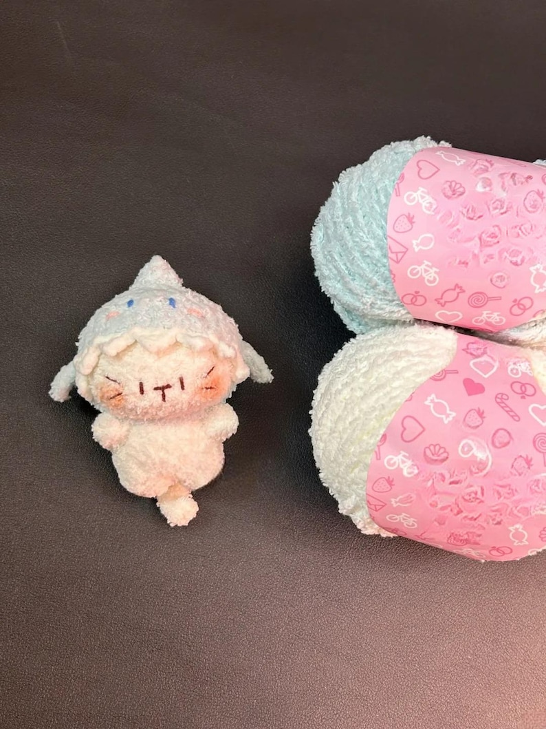 Peut inclure: Une peluche blanche avec un chapeau et des d&eacute;tails bleu clair, &agrave; c&ocirc;t&eacute; de deux pelotes de laine blanche et bleu clair. La laine a des &eacute;tiquettes roses avec de petites illustrations.