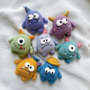 Könnte beinhalten: Eine Sammlung von sieben bunten, gehäkelten Monstermonster-Spielzeugen. Jedes Monster hat eine einzigartige Farbe, einen Gesichtsausdruck und eine Partyhütchen oder einen Hut. Die Spielzeuge sind mit einem strukturierten Stich gefertigt und haben große Wackelaugen und kleine Hörner.