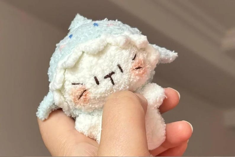 Peut inclure: Une peluche de mouton blanc avec un chapeau et des d&eacute;tails bleu clair. Le mouton a les yeux ferm&eacute;s, une petite bouche et des joues roses. Le jouet est tenu dans une main.