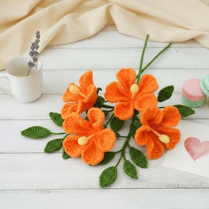 Pode incluir: Um grupo de flores de crochê laranja com folhas e caules verdes. Uma caneca branca com lavanda e uma pilha de macarons também estão presentes. Um cartão com um coração rosa está em uma superfície de madeira branca.