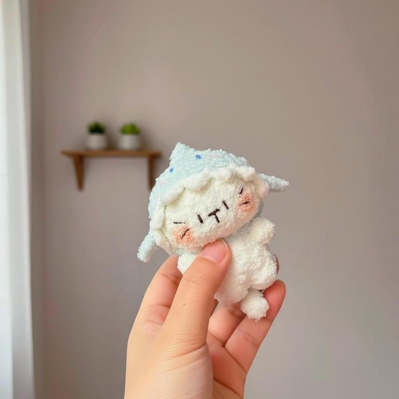 Peut inclure: Une peluche faite &agrave; la main en forme de mouton, avec un chapeau bleu clair orn&eacute; de blanc et de points bleus. Le mouton a un corps blanc, les yeux ferm&eacute;s et des joues roses. Le jouet est tenu dans une main.