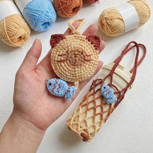 May include: Handmade crochet items including a tan cat face keychain with brown ears, a blue fish charm, and a tan and brown water bottle holder. Yarn skeins in various colors are in the background.