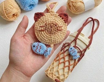 May include: A selection of handmade crochet items. A tan cat face keychain with brown ears, a blue fish charm, and a tan and brown water bottle holder. Various coloured yarn skeins are in the background.