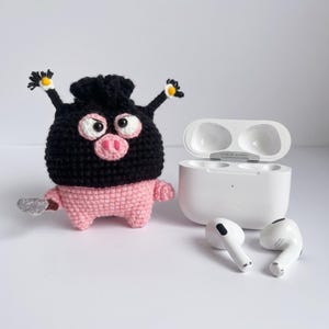 以下が含まれることがあります： 2本の触角、ピンクの鼻、小さな灰色のナイフのアクセサリーが付いた、黒とピンクの豚のようなキャラクターの編みぐるみケース。隣には、開いた状態の白いワイヤレスイヤホン充電ケースと2つの白いワイヤレスイヤホンが置かれています。