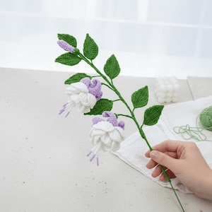 Op de afbeelding: Een handgemaakte gehaakte fuchsia bloemstengel. De stengel is groen met groene bladeren en witte en paarse bloemen. De bloem heeft lange, delicate meeldraden. De stengel wordt door een persoon vastgehouden.
