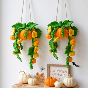May include: Two hanging crochet pumpkin decorations with green vines and yellow flowers. Below, four real pumpkins in white and orange sit on a wooden shelf next to a sign that reads Halloween Keepsakes, with a small black bat cutout on the wall.