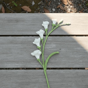 May include: A handmade crochet flower stem with white blossoms and green stem and leaves. The crochet flower is lying on a weathered wooden surface, with natural light casting shadows.