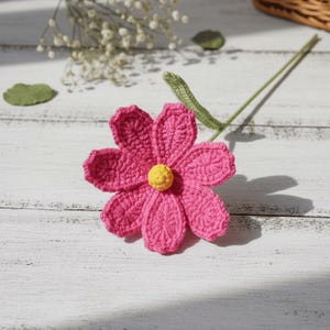 Può includere: Un fiore all'uncinetto rosa fatto a mano con un centro giallo. Il fiore ha sei petali e uno stelo verde. L'immagine mostra anche un piccolo bouquet di fiori bianchi e una foglia verde.