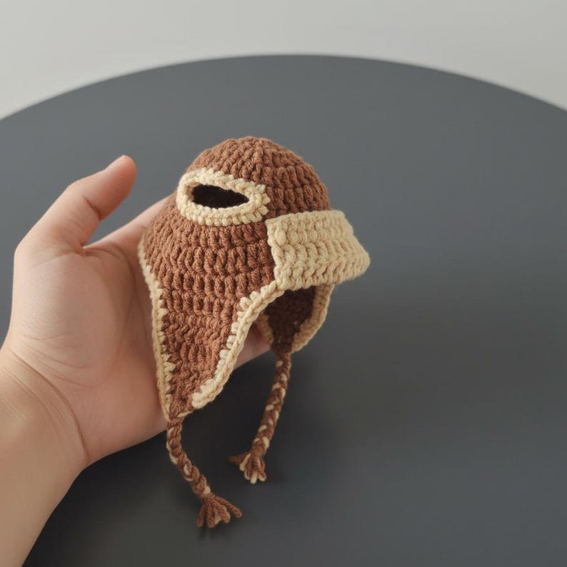 May include: A handmade crochet hat in brown and beige. The hat has ear flaps and a visor, with a small eye opening. The hat is held in a hand against a grey background. The hat has two braided tassels.