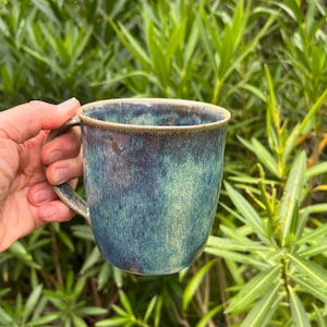 Könnte beinhalten: Ein Keramikbecher mit einer reaktiven Glasur in gemischten Schattierungen von tiefem Blau, Türkis und erdigem Braun. Der Becher hat einen abgerundeten Körper, einen breiten Rand und einen geschwungenen Henkel an der Seite mit glatter Oberfläche.