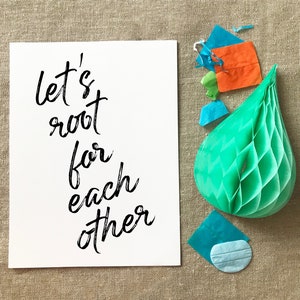 May include: A white card with black calligraphy that reads "Let's root for each other". The card is surrounded by blue and orange tissue paper and a green honeycomb ball.