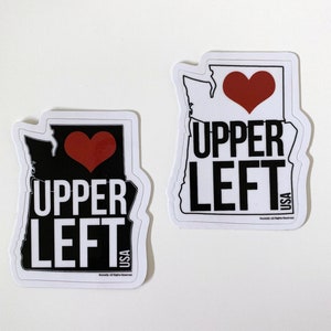 May include: Two white stickers with a black outline of the state of Oregon. The stickers have a red heart and the text "UPPER LEFT USA" in black.