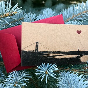 May include: A brown card with a black silhouette of the Golden Gate Bridge and a red heart-shaped balloon. The card is sitting on a bed of blue-green pine needles.