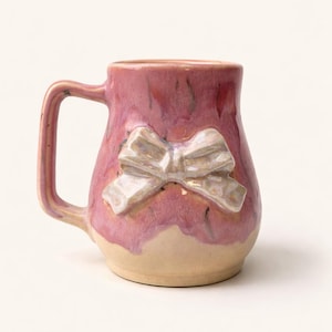 Puede incluir: Taza de cerámica hecha a mano con esmalte rosa y base color crema. La taza presenta un diseño de lazo en relieve en el centro. El asa es de color rosa a juego.