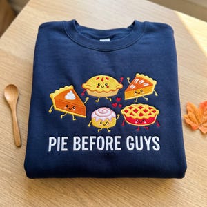 May include: A navy blue crewneck sweatshirt featuring an embroidered graphic of five smiling, anthropomorphic baked goods including pies and a cinnamon roll, with the white text "PIE BEFORE GUYS" printed below the design.
