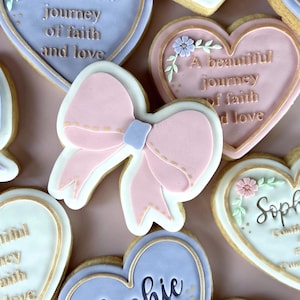 May include: Selection of pastel-coloured decorated biscuits. Heart-shaped biscuits with floral decorations and the name "Sophie" or the phrase "A beautiful journey of faith and love." Bow-shaped biscuits are also visible.