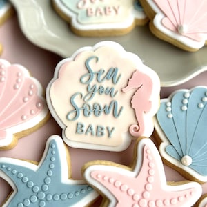 May include: A collection of sugar biscuits shaped like seashells, starfish, and seahorses, decorated with pastel pink and blue icing. Some biscuits feature the text Sea you Soon BABY in blue and pink script, accented with small white pearl details.