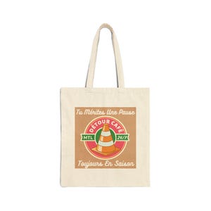 May include: A beige canvas tote bag featuring a square graphic with a traffic cone illustration in the center. The text reads Tu Mérites Une Pause, Détour Café, MTL, 24/7, and Toujours En Saison. The design uses red, green, and orange colors.
