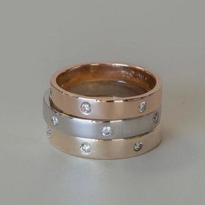 May include: Three stacked rings in rose gold, silver, and gold. Each ring features small, evenly spaced clear gemstones. The rings are displayed on a neutral background, showcasing their simple elegance and design.