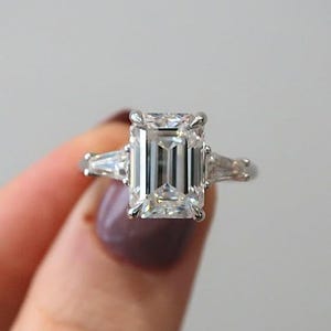 May include: A three-stone engagement ring with a large emerald-cut center diamond and two smaller, tapered baguette diamonds on either side. The ring is set in a silver band and is held up by a person's hand.