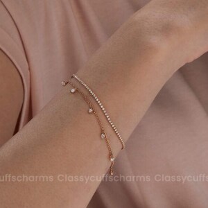 Könnte beinhalten: Armband-Set in Roségold. Ein Armband hat eine zarte Kette mit kleinen, hängenden Anhängern. Das andere ist eine dünne, funkelnde Linie aus kleinen Steinen. Die Armbänder werden am Handgelenk getragen.