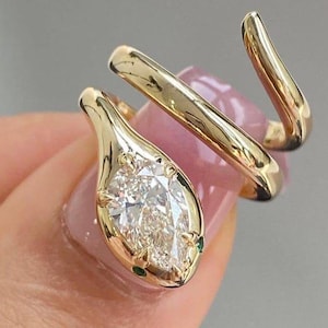 May include: A gold-colored ring in the shape of a snake, with a pear-shaped diamond as the head. The snake's body wraps around the finger. Small green stones accent the head. The ring is shown on a person's finger.