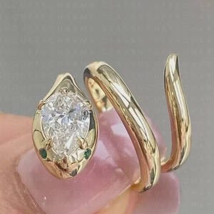 May include: A gold bypass ring featuring a pear-shaped clear gemstone at one end and a smooth, tapered gold band that wraps around the finger. The setting includes two tiny green accent stones positioned at the base of the pear-shaped gem.
