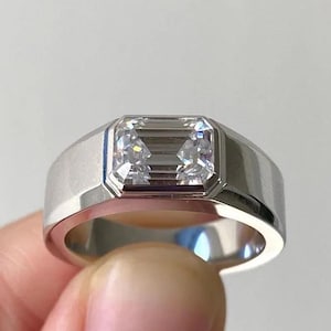 May include: A silver ring with a rectangular, clear gemstone. The ring has a wide band and a modern, minimalist design. The gemstone is set in a bezel setting, and the ring is held by a person's finger.