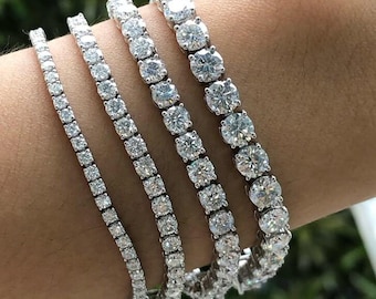 May include: Four diamond tennis bracelets of varying widths are displayed on a wrist. The bracelets feature round-cut, clear gemstones set in a silver or white metal. The bracelets are arranged from narrowest to widest, showcasing the different sizes.