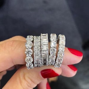 Può includere: Una collezione di sei anelli eternity di diamanti, ognuno con un taglio diverso di diamanti bianchi scintillanti. Gli anelli sono mostrati su una mano con smalto rosso, mettendo in risalto i vari design.