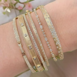 Puede incluir: Varios brazaletes dorados de diferentes diseños y anchos. Algunos están adornados con pequeños diamantes, otros tienen una superficie texturizada. Los brazaletes están apilados, creando un efecto de capas. El estilo general es elegante y lujoso.