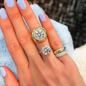 Op de afbeelding: Een hand toont verschillende gouden ringen met heldere edelstenen. Eén ring heeft een grote, ronde diamant omringd door kleinere stenen. Een andere ring heeft een enkele, prominente diamant. Een derde ring is een eenvoudige gouden band.