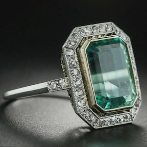 Puede incluir: Un anillo de estilo vintage con una gran gema verde rectangular con talla esmeralda. La gema está rodeada de pequeñas piedras claras en un engaste de metal plateado. El anillo tiene un diseño clásico y elegante.