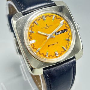 May include: A vintage Ulysse Nardin automatic wristwatch with a square silver case and a bright orange dial. The watch features a black leather band and displays the day and date. The dial has luminous markers and hands.