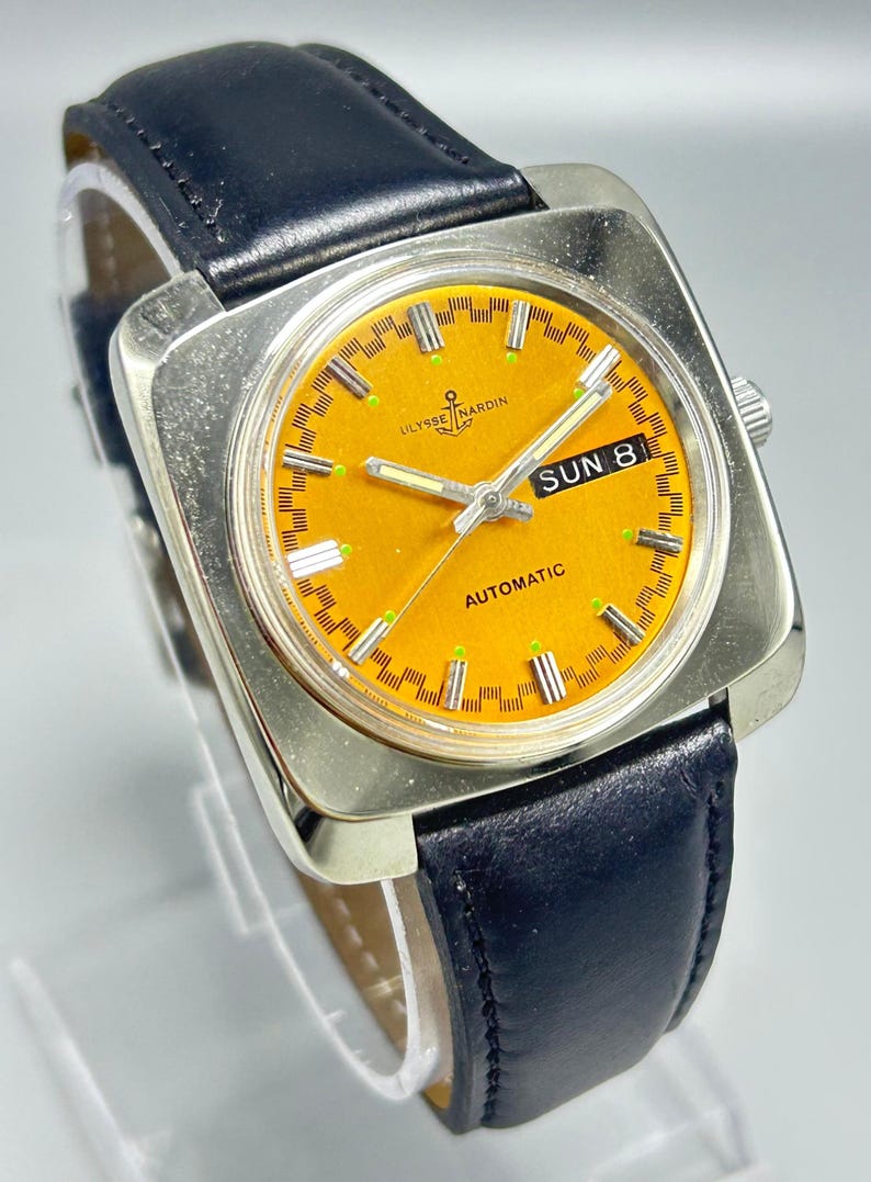 May include: A vintage Ulysse Nardin automatic wristwatch with a square silver case and a bright orange dial. The watch features a black leather band and displays the day and date. The dial has luminous markers and hands.
