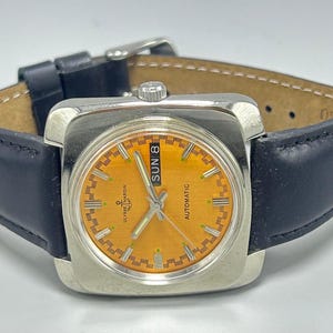 May include: A vintage Ulysse Nardin automatic watch with a square silver case and a bright orange dial. The watch features a black leather strap and displays the day of the week. The watch face has silver hands and markers.