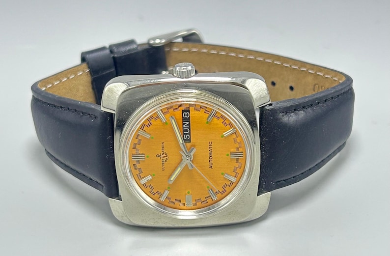 May include: A vintage Ulysse Nardin automatic watch with a square silver case and a bright orange dial. The watch features a black leather strap and displays the day of the week. The watch face has silver hands and markers.