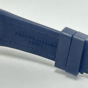 May include: Close-up of a dark blue watch strap with the brand name "FRANCK MULLER GENEVE" embossed in white. The strap is made of a flexible material and has a buckle.