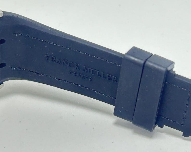 May include: Close-up of a dark blue watch strap with the brand name "FRANCK MULLER GENEVE" embossed in white. The strap is made of a flexible material and has a buckle.