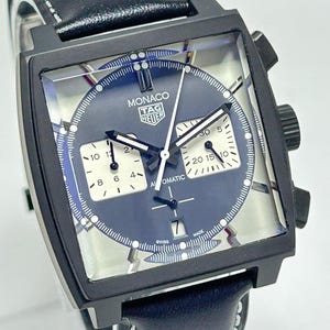 Puede incluir: Un reloj automático Monaco Tag Heuer negro con esfera cuadrada y esfera azul oscuro. El reloj presenta subesferas, agujas e indicadores blancos, y una correa de cuero negro con costuras blancas. La esfera del reloj tiene el texto "MONACO TAG HEUER AUTOMATIC SWISS MADE".