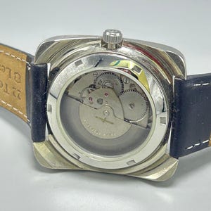 May include: A silver-toned, square-faced wristwatch with a clear back, showcasing the intricate inner workings of the watch. The watch has a black leather strap and a crown at the top. The watch mechanism is visible through the clear back.