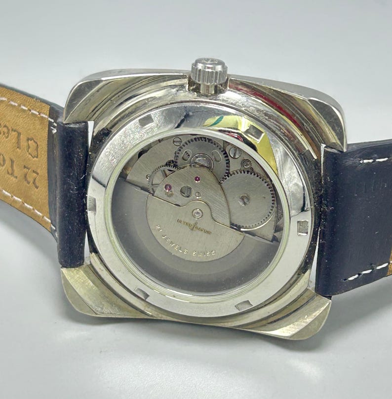 May include: A silver-toned, square-faced wristwatch with a clear back, showcasing the intricate inner workings of the watch. The watch has a black leather strap and a crown at the top. The watch mechanism is visible through the clear back.